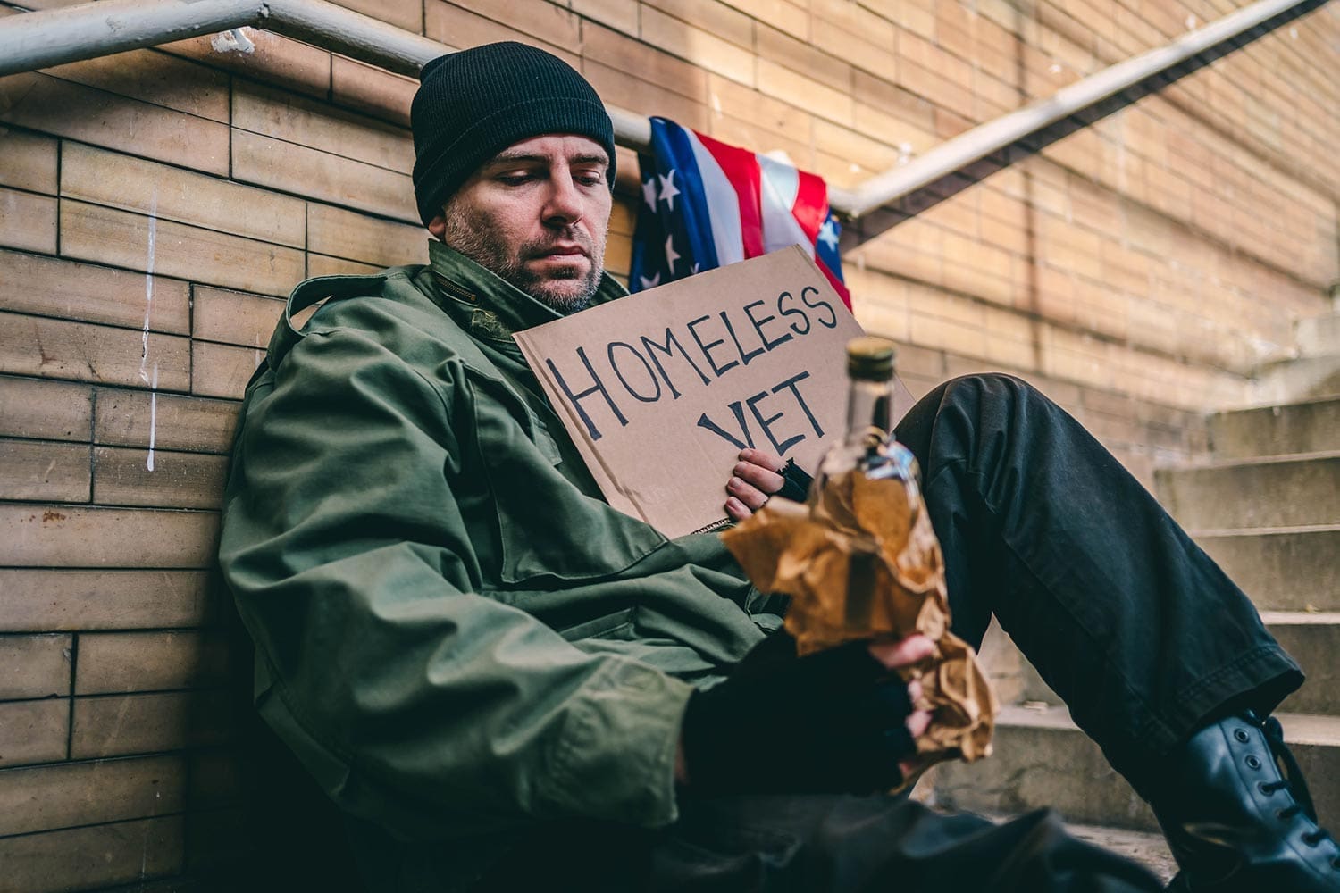 Featured image for VA Homeless Program, showing a homeless Veteran sitting on the stairs with a bottle of alcohol in one hand and holding up a sign in his other hand